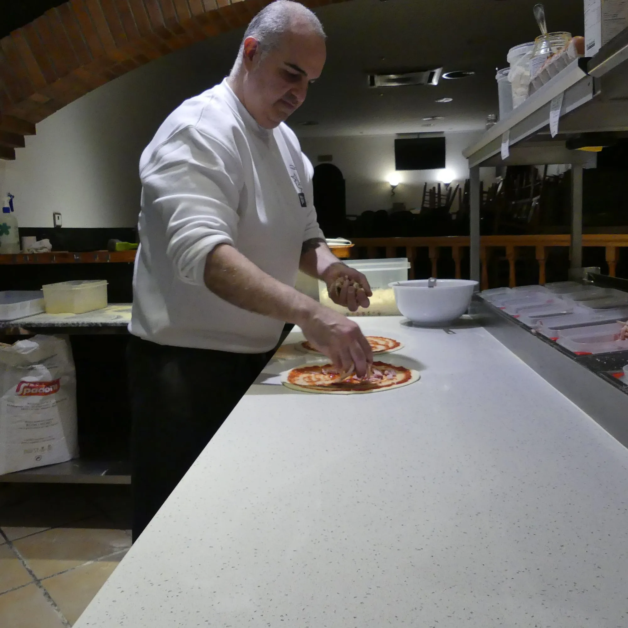 Luiggi extendiendo ingredientes sobre una base de pizza a mano en el obrador de la Trattoria (2020)