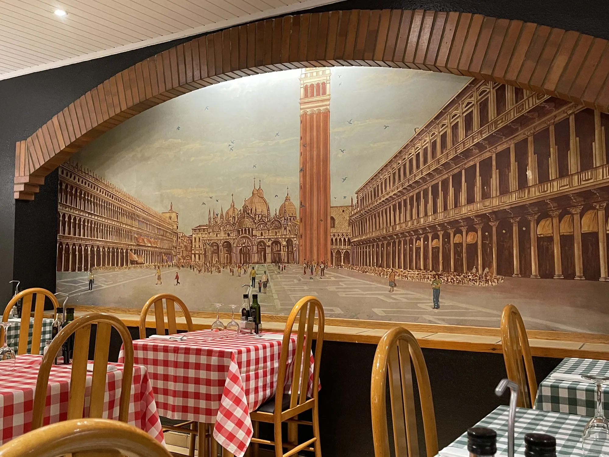 Sala de la Trattoria da Luiggi, en Olot, con el mural de la plaza de San Marcos y mesas de cuadros