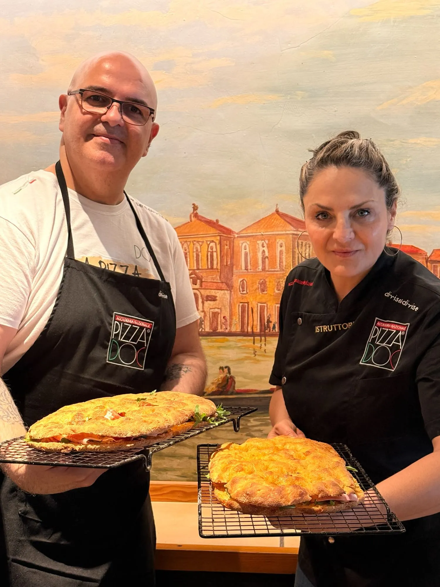 Luiggi y Patrizia Di Leo sonriendo con una pizza cada uno en bandeja, en el curso de junio de 2026 en la Trattoria