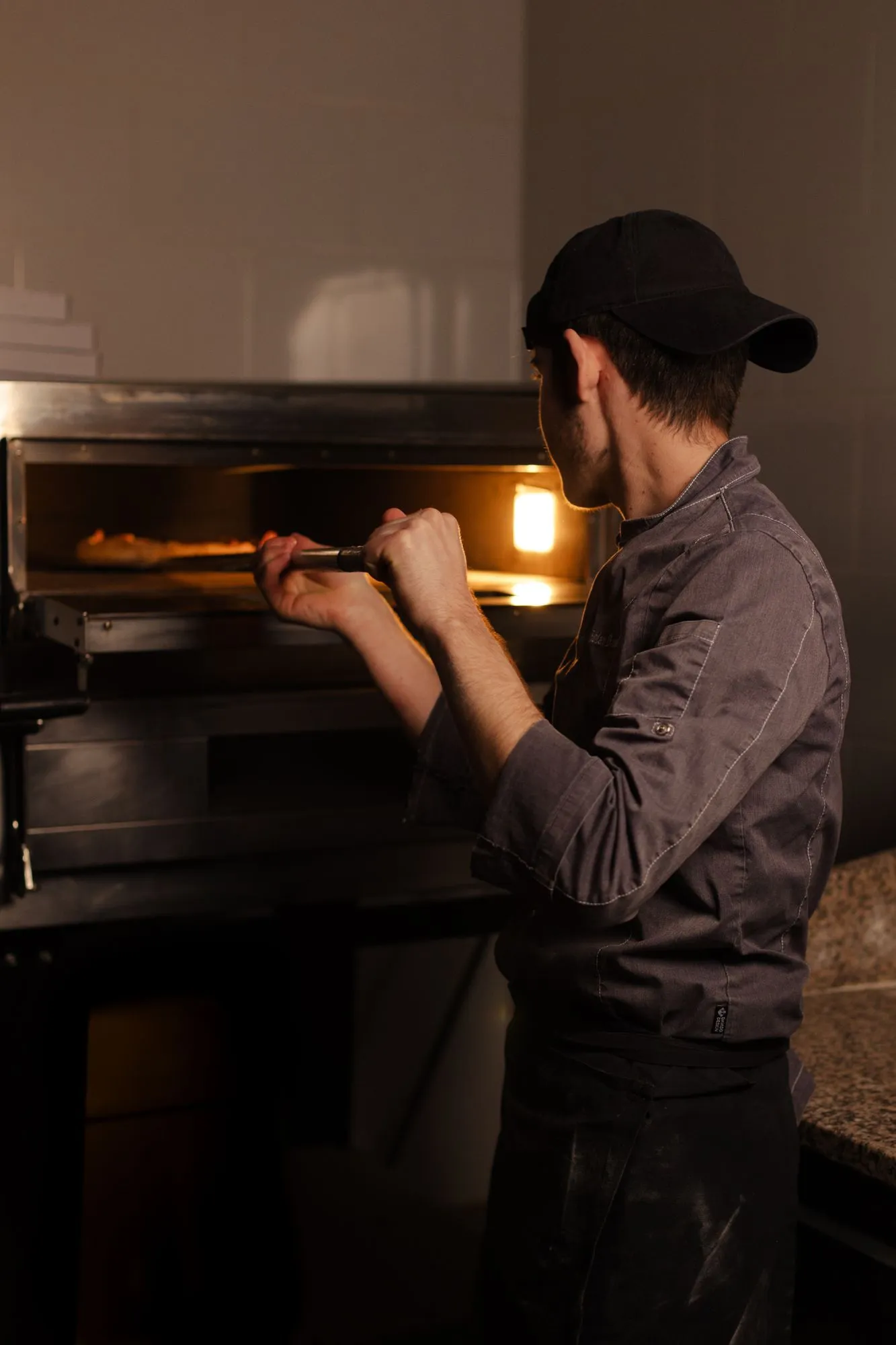 Pizzero con gorra y chaquetilla gris metiendo una pizza en un horno eléctrico de pizzería, de espaldas y a media luz
